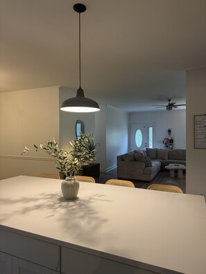 Een moderne keuken met een centraal eiland, een eetgedeelte met rieten stoelen, en een woonkamer met een bank en een plafondventilator.