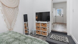 Un dormitorio con cama, televisor, cajas de almacenamiento, un colgador de macramé y una puerta que da a otra habitación.