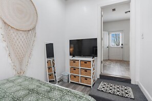 Een slaapkamer met een bed, een televisie, opbergkisten, een macramé wanddecoratie en een deur naar een andere kamer.