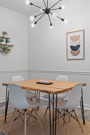 Een eetruimte met een houten tafel, witte stoelen, een moderne kroonluchter, een lijst met abstracte kunst en een plantenplank.