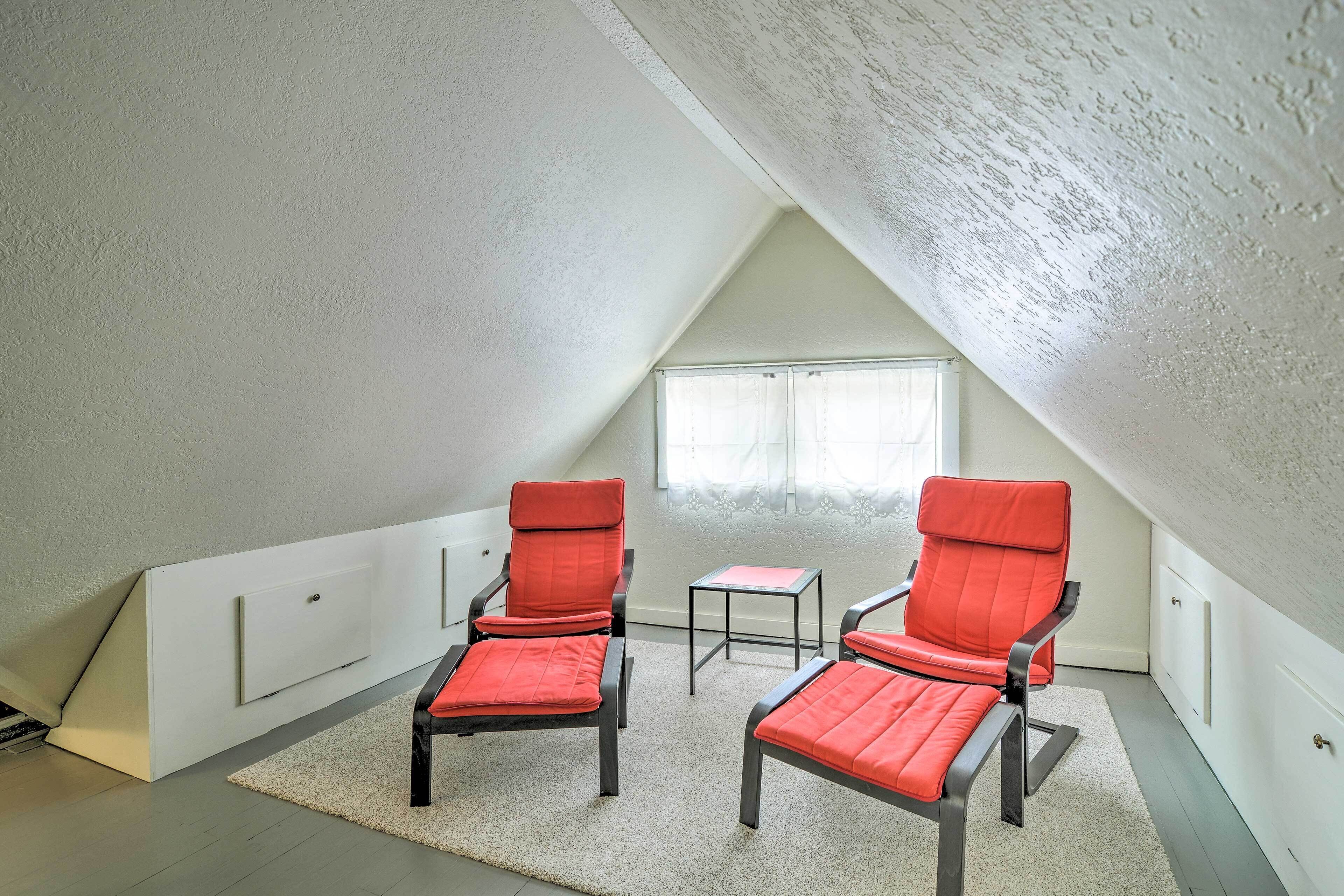A small, triangular attic room with two red chairs, a small table, and a window.