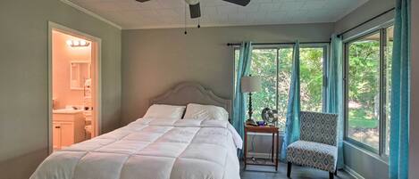 A bedroom with a bed, a chair, a nightstand, a ceiling fan, and large windows with curtains.