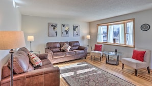 A living room with a brown leather sofa, a glass coffee table, and a red chair. There are three wall-mounted lamps and a large window with curtains. - Rapid City Home - Walk to Canyon Lake Park (Rapid City)