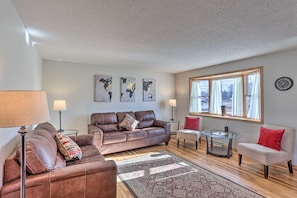 A living room with a brown leather sofa, a glass coffee table, and a red chair. There are three wall-mounted lamps and a large window with curtains. - Rapid City Home - Walk to Canyon Lake Park (Rapid City)