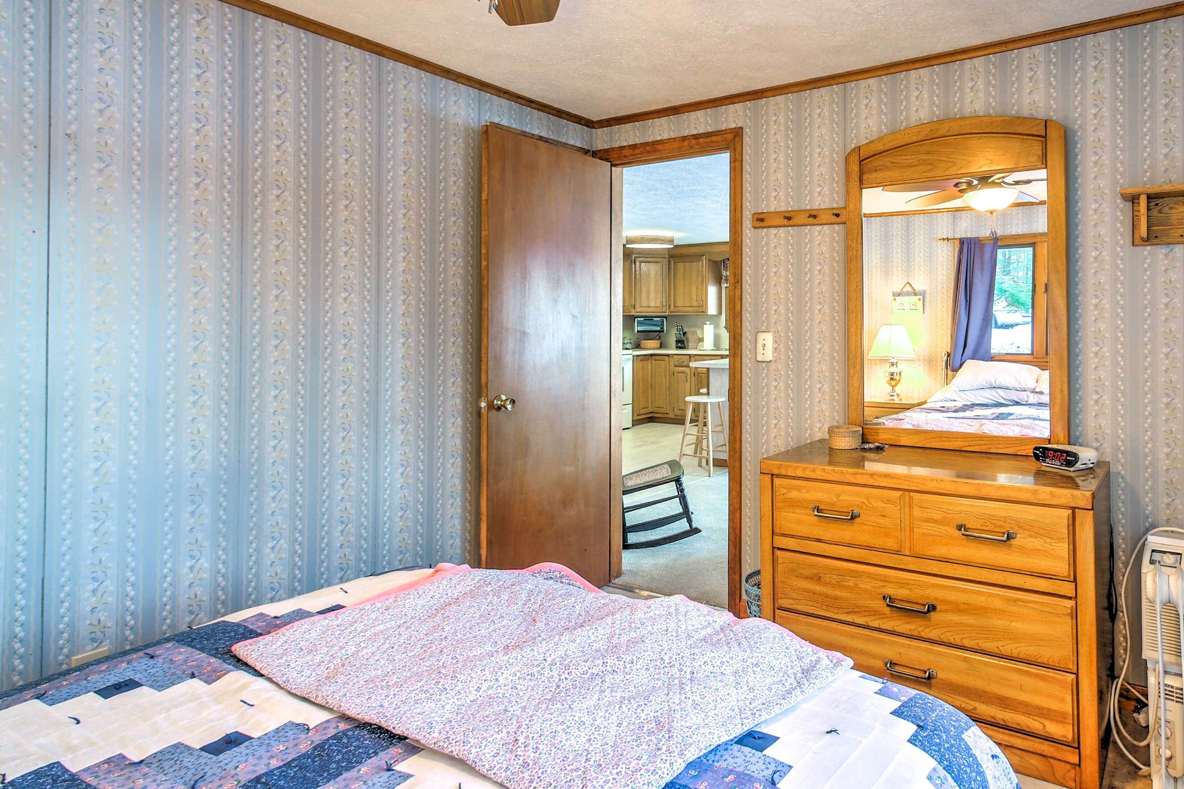 A bedroom with a bed, a dresser, a mirror, and a door leading to a kitchen area.