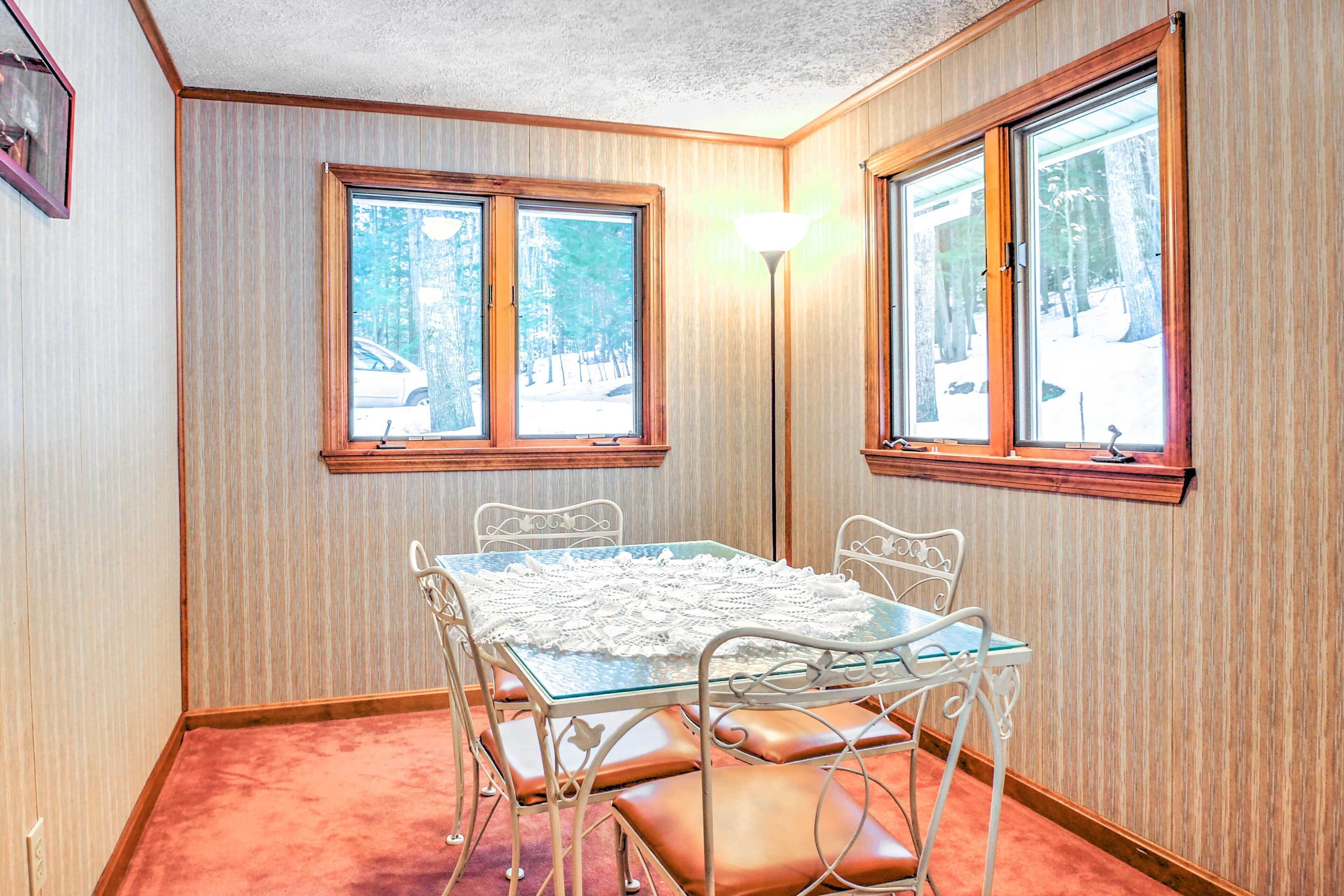 Una habitación de esquina con una mesa de vidrio, sillas de metal blanco y un banco. Se observan dos ventanas con marcos de madera, y una lámpara de pie.