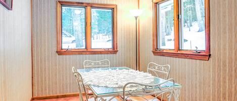 Una habitación de esquina con una mesa de vidrio, sillas de metal blanco y un banco. Se observan dos ventanas con marcos de madera, y una lámpara de pie.
