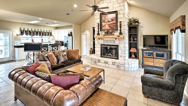 A spacious living room with a stone fireplace, leather furniture, a television, and a kitchen in the background.
