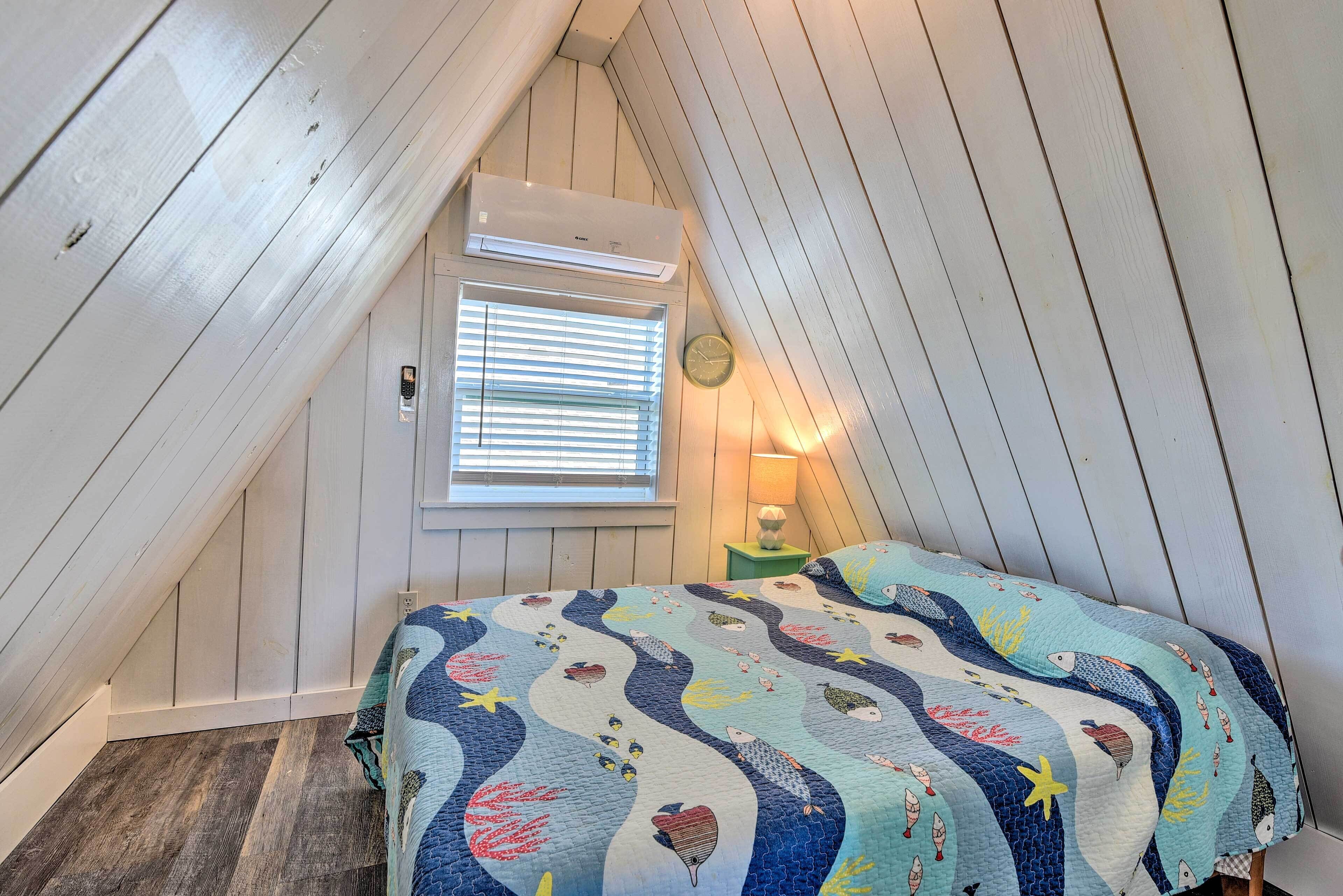 Un dormitorio con techo inclinado, una ventana con persianas, una cama con colcha, una lámpara de noche y un reloj de pared.