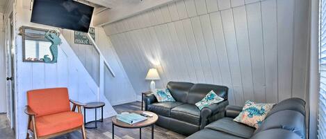 A cozy living room with a sofa, armchair, and a small table. A ceiling fan and a flat-screen TV are mounted on the wall.