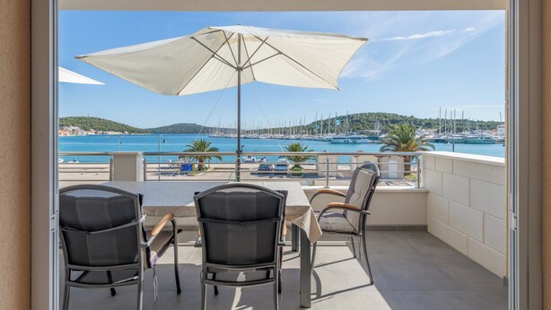 A balcony with a table and chairs, a large umbrella, and a view of a marina with boats and hills in the distance. - Seafront apt With Terrace, 4 Bedrooms and Mooring (Rogoznica)
