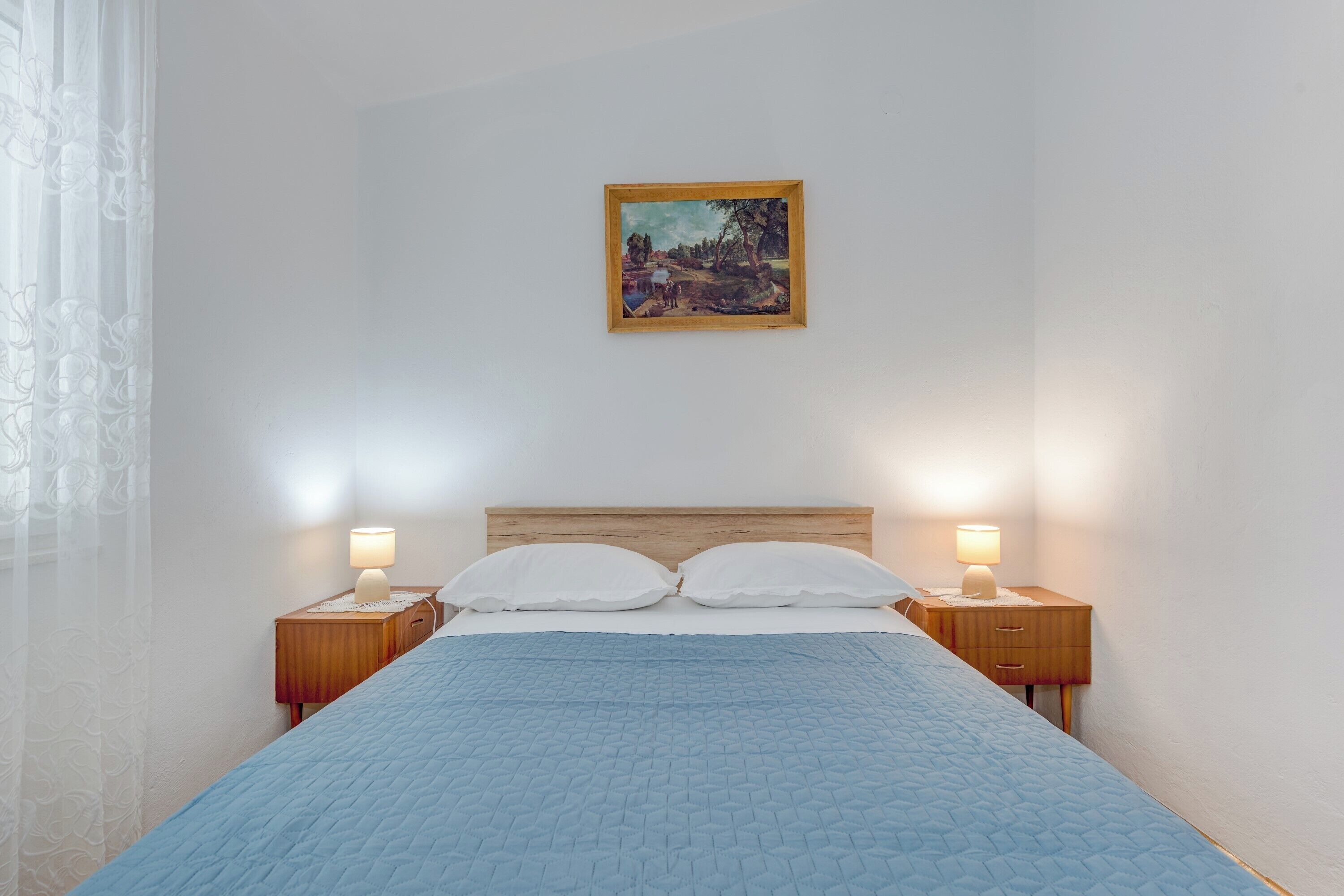 Una cama bien hecha con colcha azul, dos lámparas de noche y un cuadro en la pared.