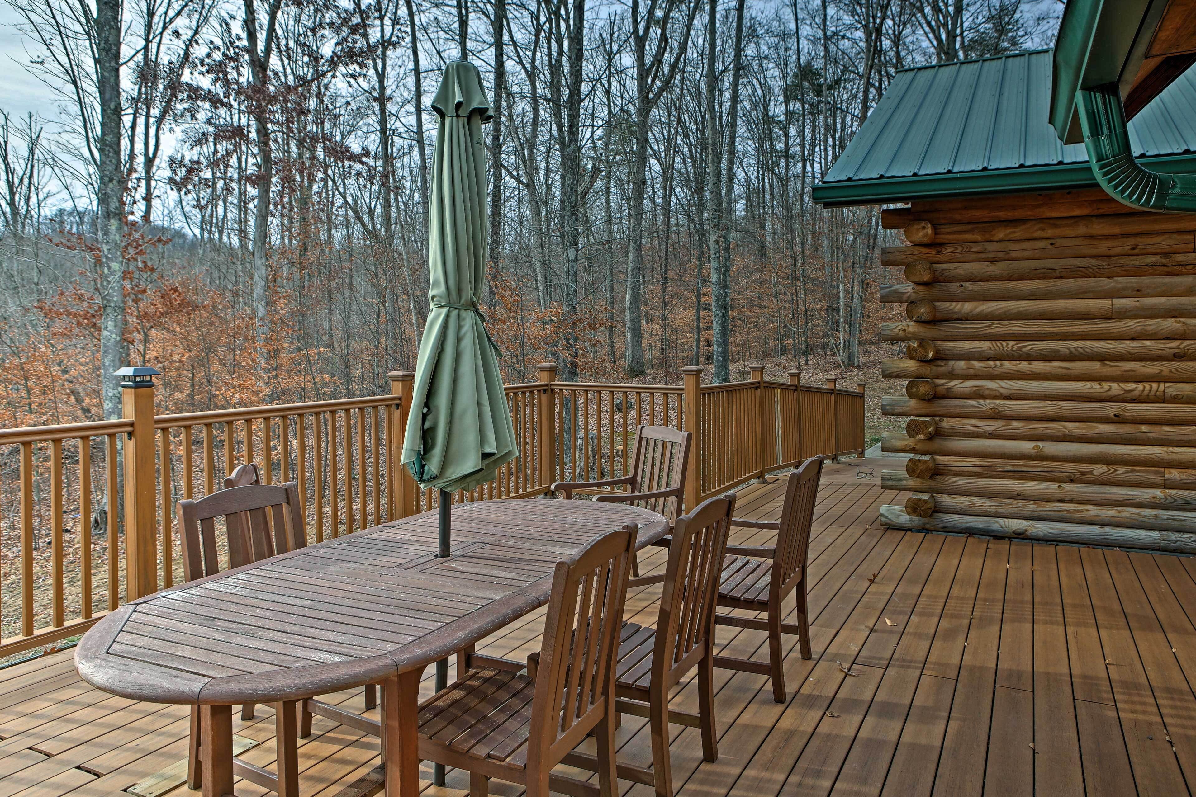 A wooden deck with a table and chairs, surrounded by a railing, overlooking a forested area.