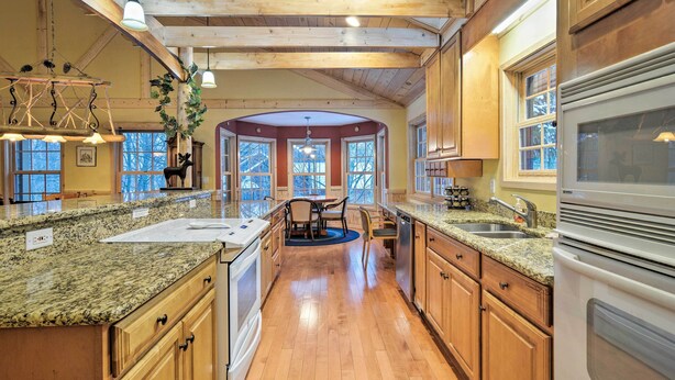A spacious kitchen with granite countertops, wooden cabinets, and a large island. There is a dining area with a table and chairs, and a view of the outdoors through large windows. - 2 Mi to Schuss Mtn! Northern Retreat w/ Game Room (Bellaire)