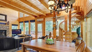 A wooden cabin interior with a dining table, chairs, a fireplace, a television, and a spiral staircase.