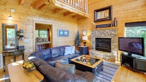 A cozy log cabin interior with a large leather sofa, a stone fireplace, a television, and a wooden balcony.