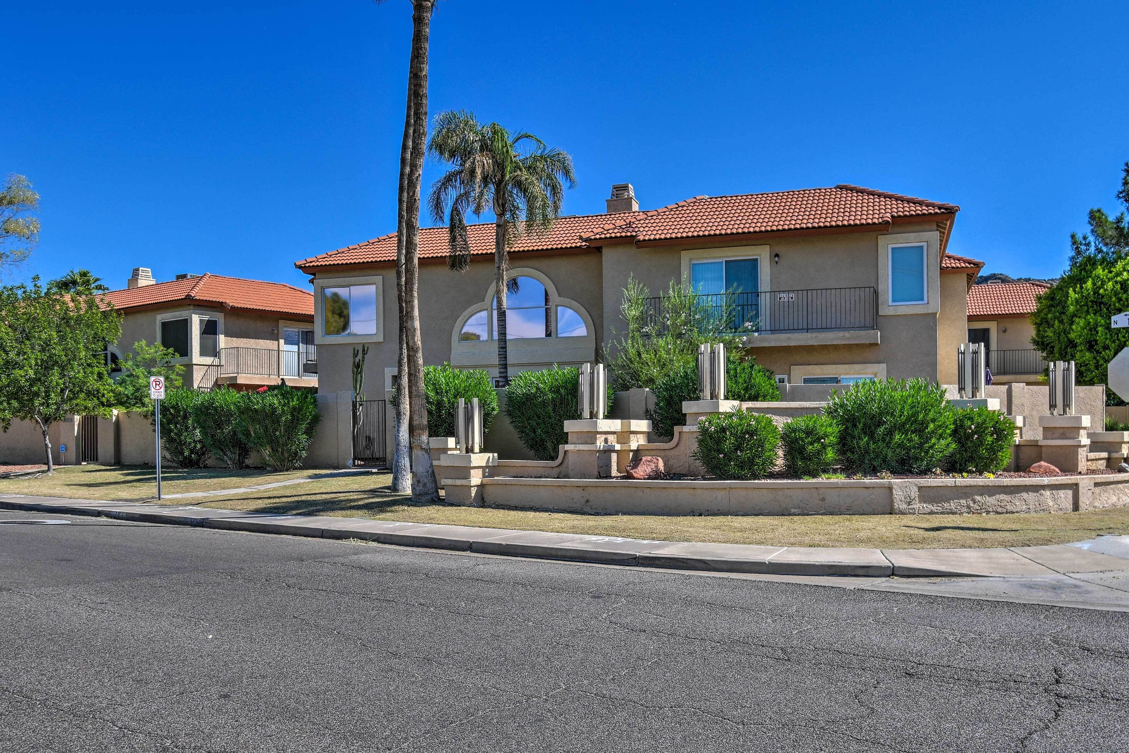 A residential building with a red-tiled roof, multiple balconies, and a landscaped front yard with palm trees and shrubs.
