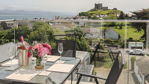 A table set with a champagne bottle, glasses, and pink flowers, overlooking a scenic view of a hill and sea. - Golygfa'r Gastell / Castle View (Criccieth)