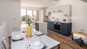 A modern kitchen with white cabinets, a built-in oven, and a microwave. There is a dining area with a table set for two, and a large window providing a view of the outdoors. - Golygfa'r Gastell / Castle View (Criccieth)