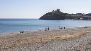 A pebbled beach with a castle on a hill, clear blue sea, and houses in the background. - Golygfa'r Gastell / Castle View (Criccieth)