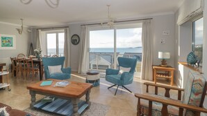 Ein geräumiges Wohnzimmer mit einem großen Fenster und Meerblick. Zwei blaue Sessel, ein Holztisch und ein Essbereich mit Holztischen und Stühlen.