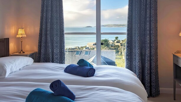 Ein Hotelzimmer mit einem großen Bett, zwei Kissen, einem Nachttisch mit Lampe und Blick auf das Meer und Gebäude.
