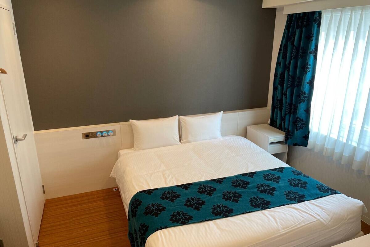 A hotel room with a bed, a bedside table, a nightstand, and a window with curtains.