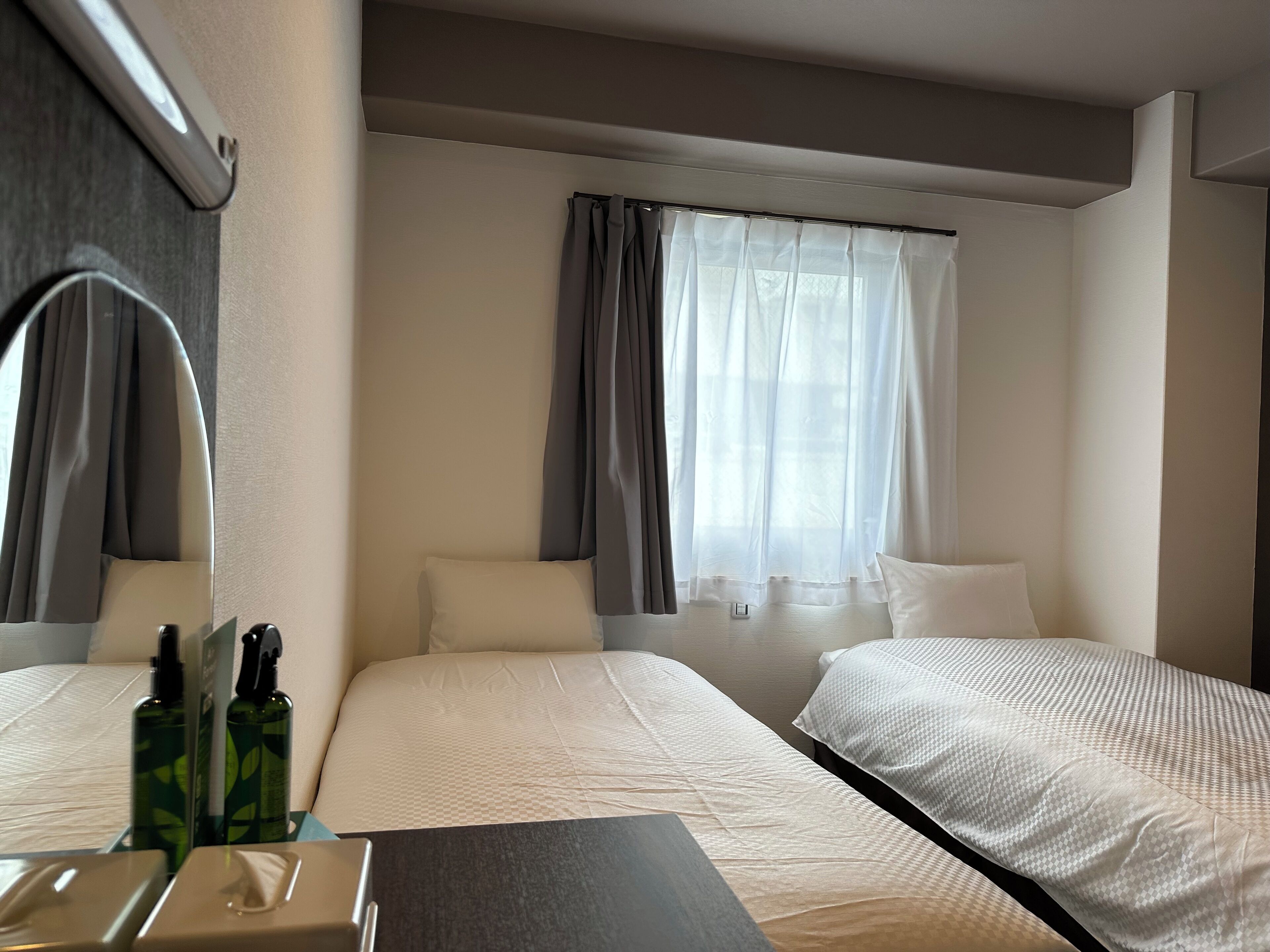 Habitación de hotel con dos camas, un espejo grande, una mesita de noche con artículos de aseo y una ventana con cortinas.