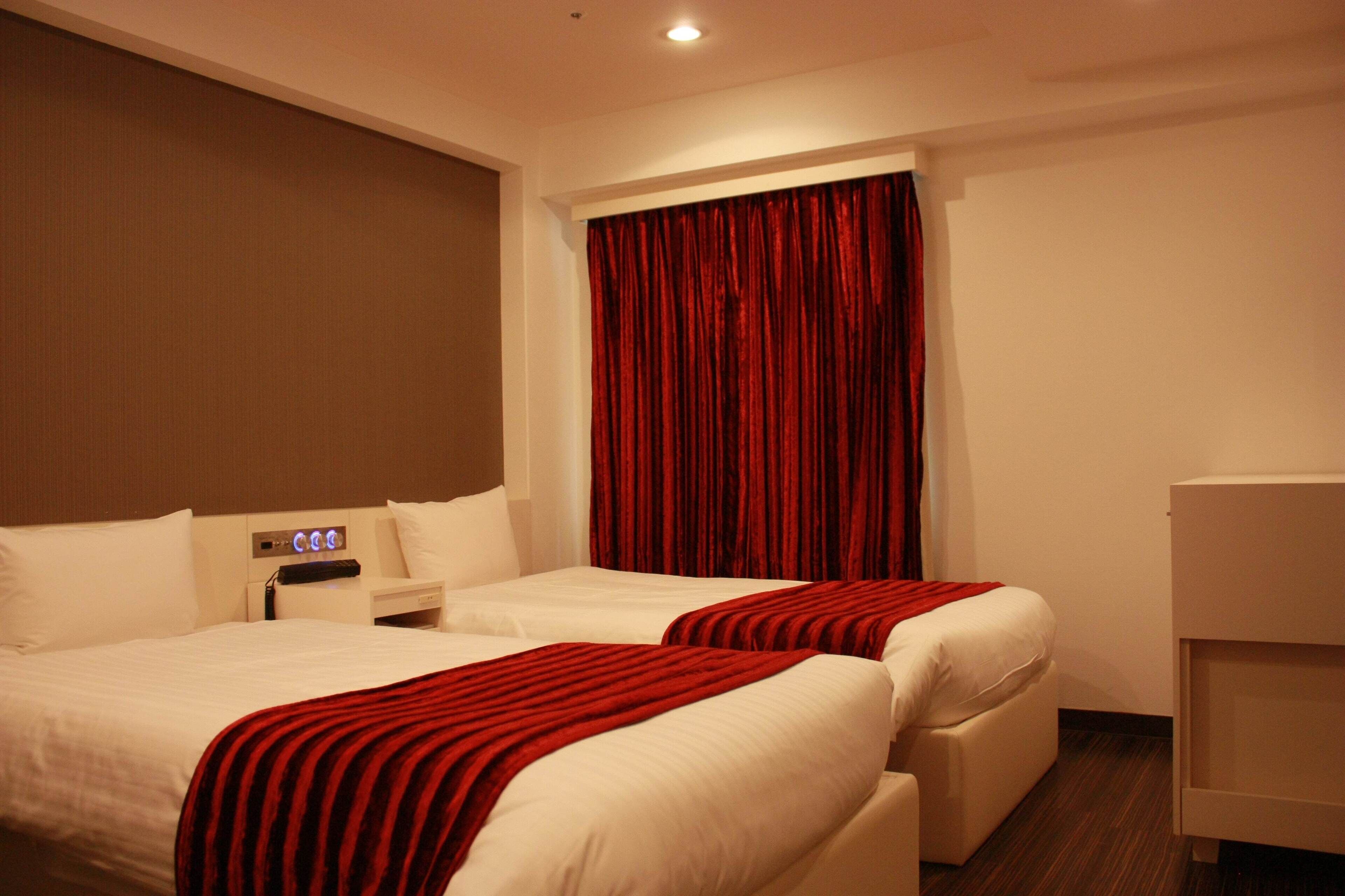 Ein Hotelzimmer mit zwei Betten, einem Nachttisch, einer Bedienfeld und einem Fenster mit roten Vorhängen.