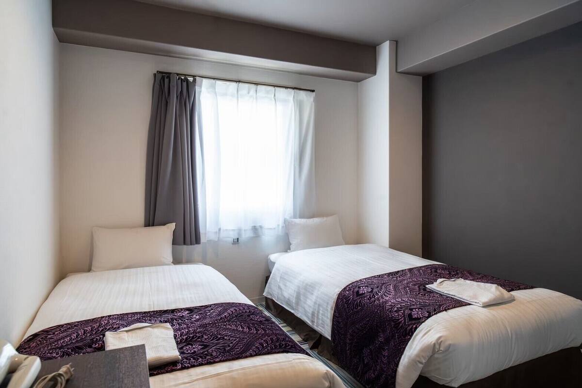 A hotel room with two beds, a window with curtains, and a desk with a lamp.