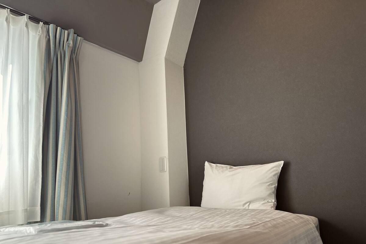 A hotel room with a bed, a window with curtains, and a plain wall.