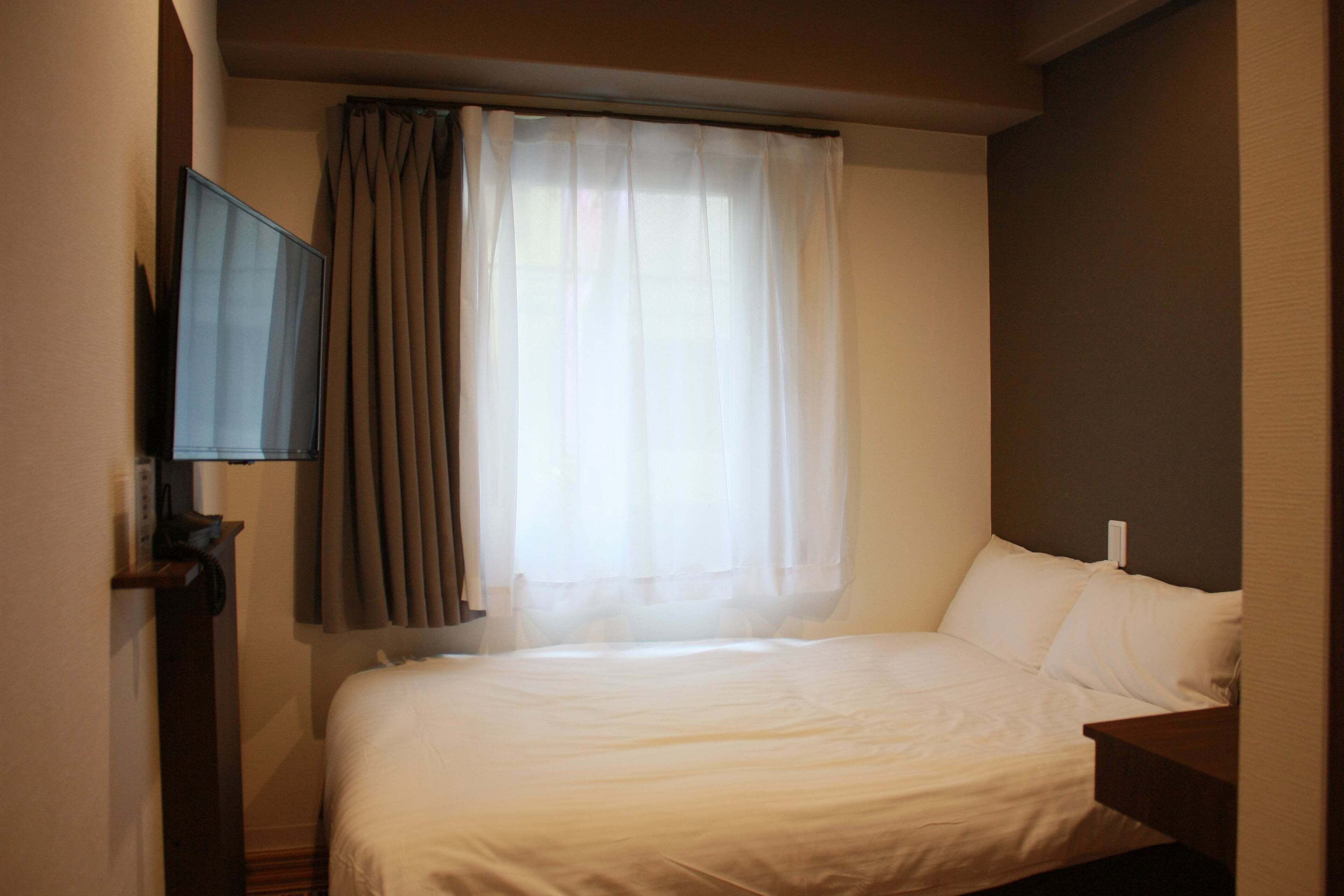 Quarto de hotel com uma cama, televisão de tela plana na parede e janela com cortinas.