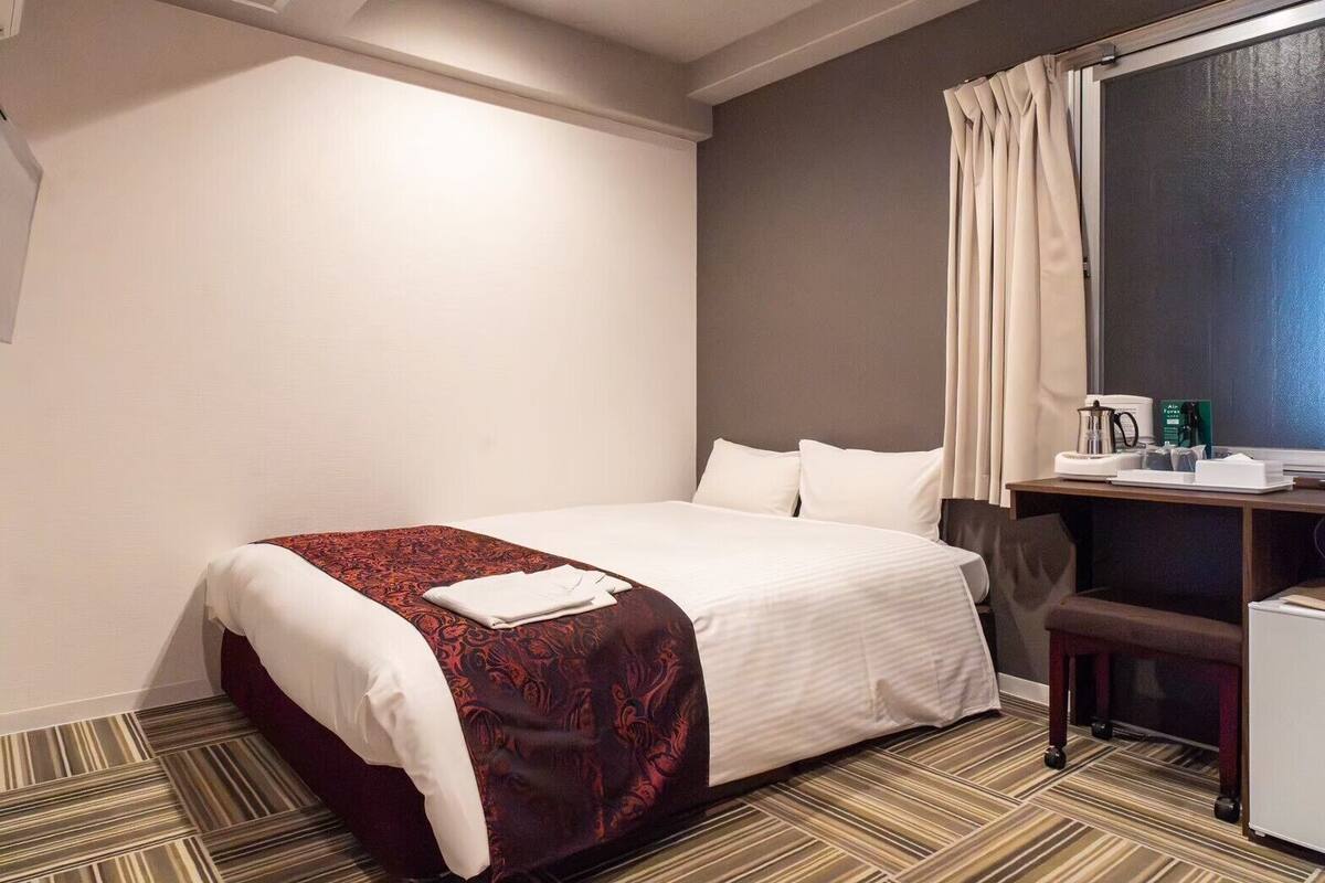 A hotel room with a bed, a desk with a chair, a coffee maker, and a window with curtains.