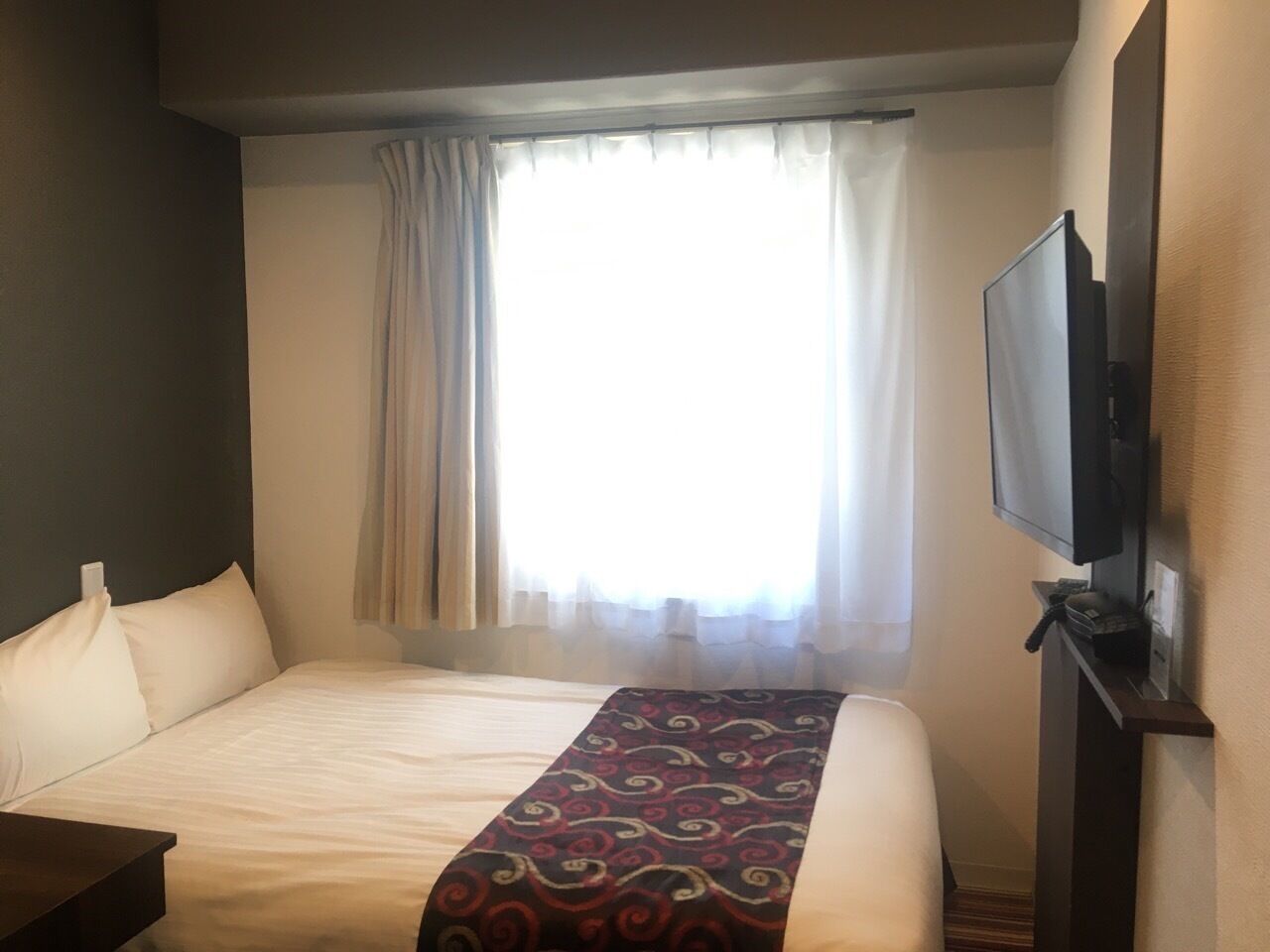 A hotel room with a bed, a flat-screen TV mounted on the wall, and a window with curtains.