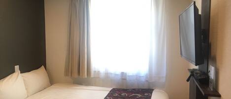 A hotel room with a bed, a flat-screen TV mounted on the wall, and a window with curtains.