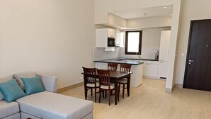Una sala de estar moderna con un sofá gris, una mesa de comedor de madera y sillas. Se ve una cocina al fondo con armarios blancos y un microondas negro.