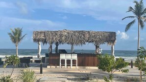 Un restaurante frente a la playa con techo de paja, mesas al aire libre y vistas al océano.