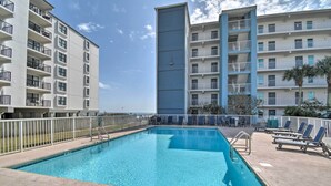 Une piscine entourée d’une clôture blanche et de chaises longues, bordée d’immeubles à plusieurs étages dotés de balcons, le tout sous un ciel dégagé.