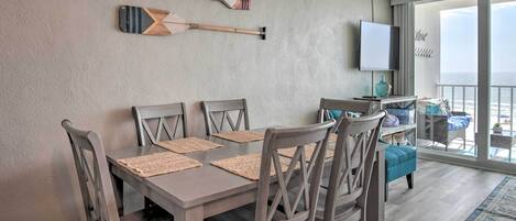 A dining area with a table and chairs, a TV mounted on the wall, and a view of the ocean through large windows.