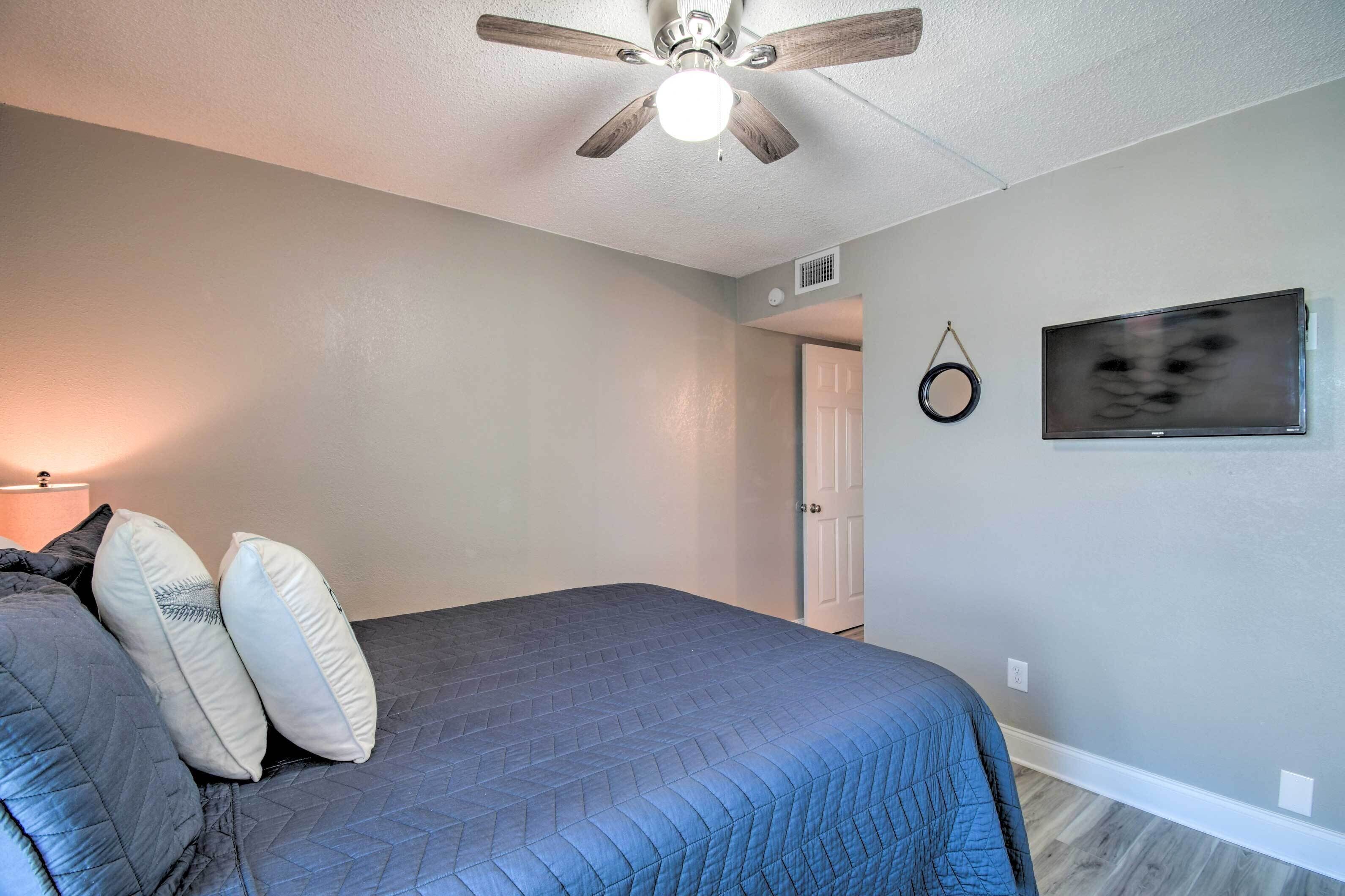 Une chambre à coucher avec un lit, des oreillers, un ventilateur de plafond, un téléviseur fixé au mur et une porte.