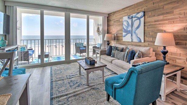 Un vaste espace de vie avec une grande porte-fenêtre coulissante donnant sur un balcon avec vue sur l’océan. On y trouve un canapé blanc, un fauteuil bleu et une petite table basse agrémentée d’un vase décoratif. Un panneau mural en bois et une œuvre d’art abstraite sont visibles.
