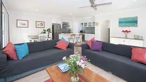 A modern living room with a dark grey sofa, colorful cushions, a wooden coffee table with a book and flowers, and a kitchen in the background. - LUXURY HAVEN! Beautiful, central, wifi & spacious! Walking to everything! (Yamba)