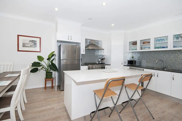 A modern kitchen with white cabinetry, a stainless steel refrigerator, a gas stove, and a microwave. There is a dining area with a table and chairs, and a potted plant.