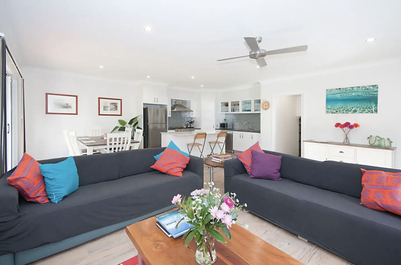 A modern living room with a dark grey sofa, colorful cushions, a wooden coffee table with a book and flowers, and a kitchen in the background.