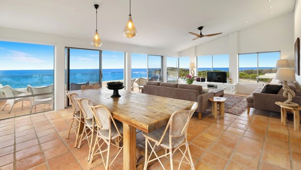Un amplio salón con una gran mesa de madera y sillas de mimbre, un sofá y un televisor. La habitación tiene ventanas grandes con vistas al océano.