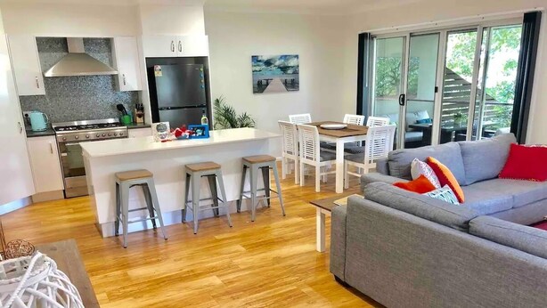 Et moderne køkken med en central ø, et spiseområde med hvide stole og en grå sofa med røde puder.