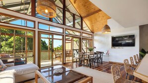 A spacious living area with large windows, a glass coffee table, wicker furniture, and a flat-screen TV mounted on the wall.