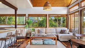 A spacious living area with a large sofa, a coffee table, and a dining area with a wooden table and chairs. There are large windows providing views of greenery outside.
