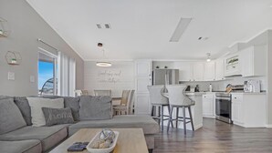 Ein modernes Wohnzimmer mit grauem Sofa, einem Holztisch und einer Küchenzeile mit weißen Schränken und einem Edelstahl-Kühlschrank.