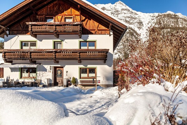 A chalet with a wooden balcony, a bench outside, and a snow-covered mountain in the background.